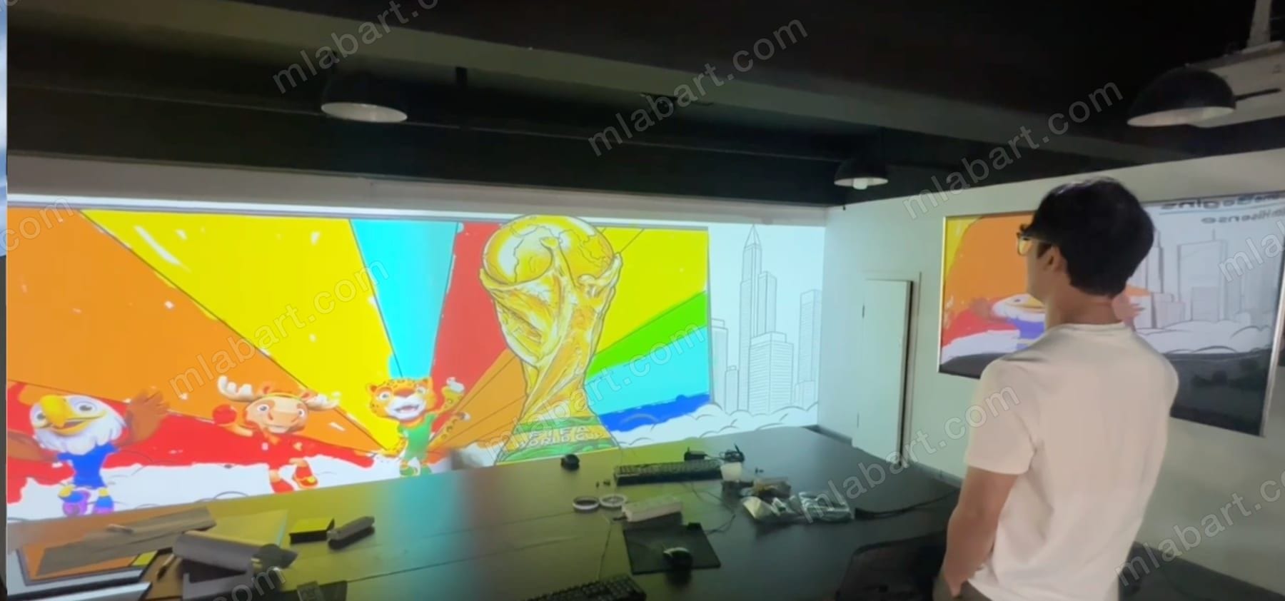 The graffiti wall content under test on a full-width wall in the MLABART facility in China before shipping, with workstations and keyboards along the bench and a second screen mirroring the canvas